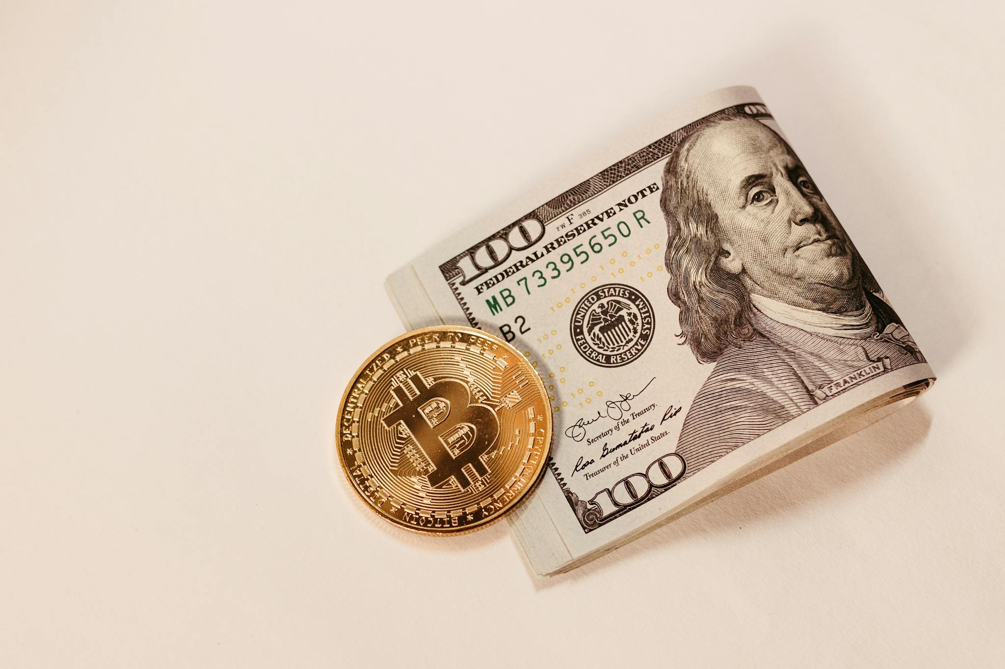 A Bitcoin coin placed next to a rolled US hundred dollar bill, symbolizing cryptocurrency and traditional currency.