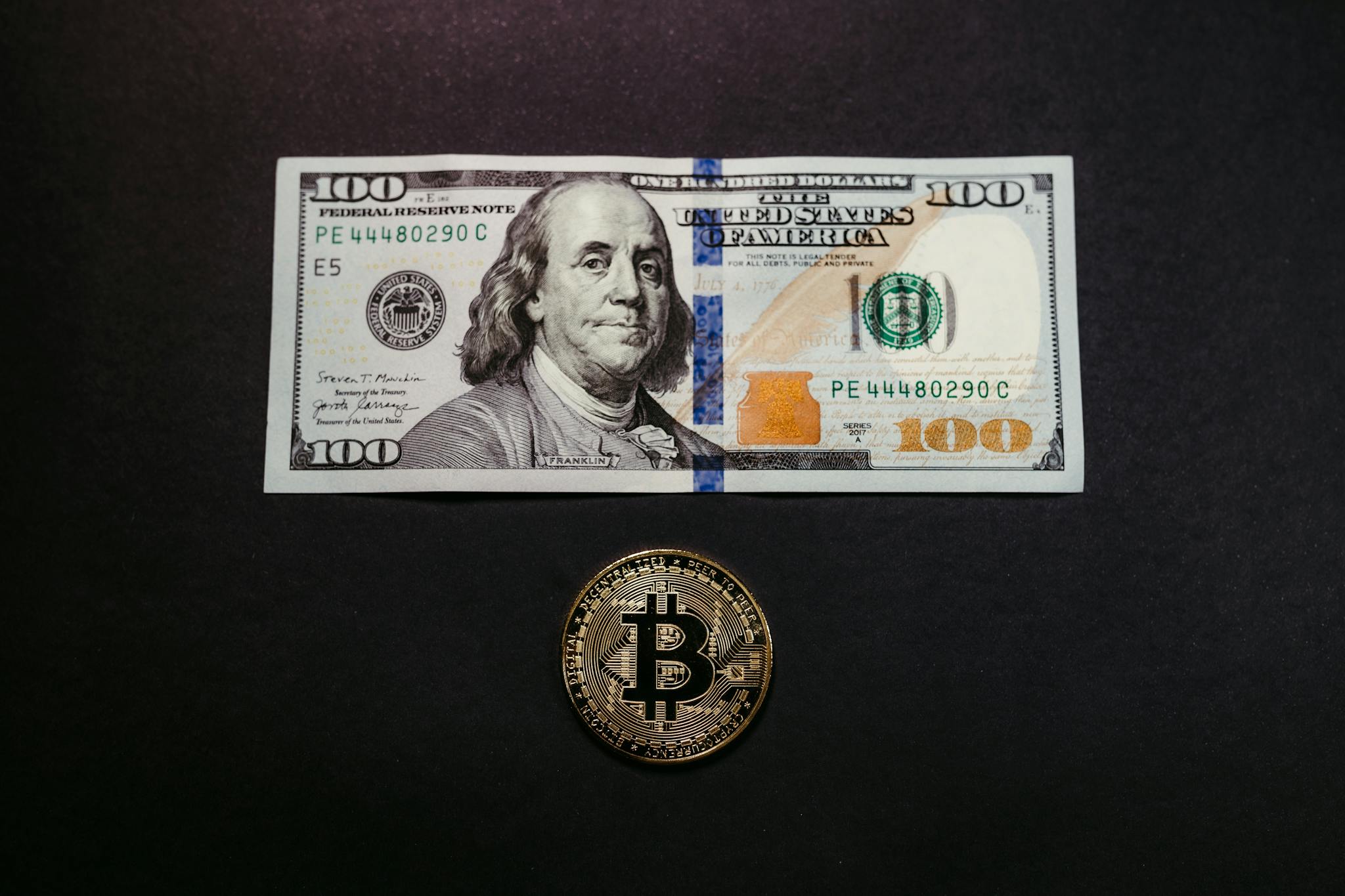 Image of a US 100 dollar bill and a Bitcoin coin on a black background, symbolizing traditional and digital currency.